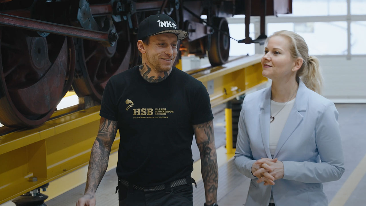 A woman and a man in a workshop, with a steam locomotive in the background