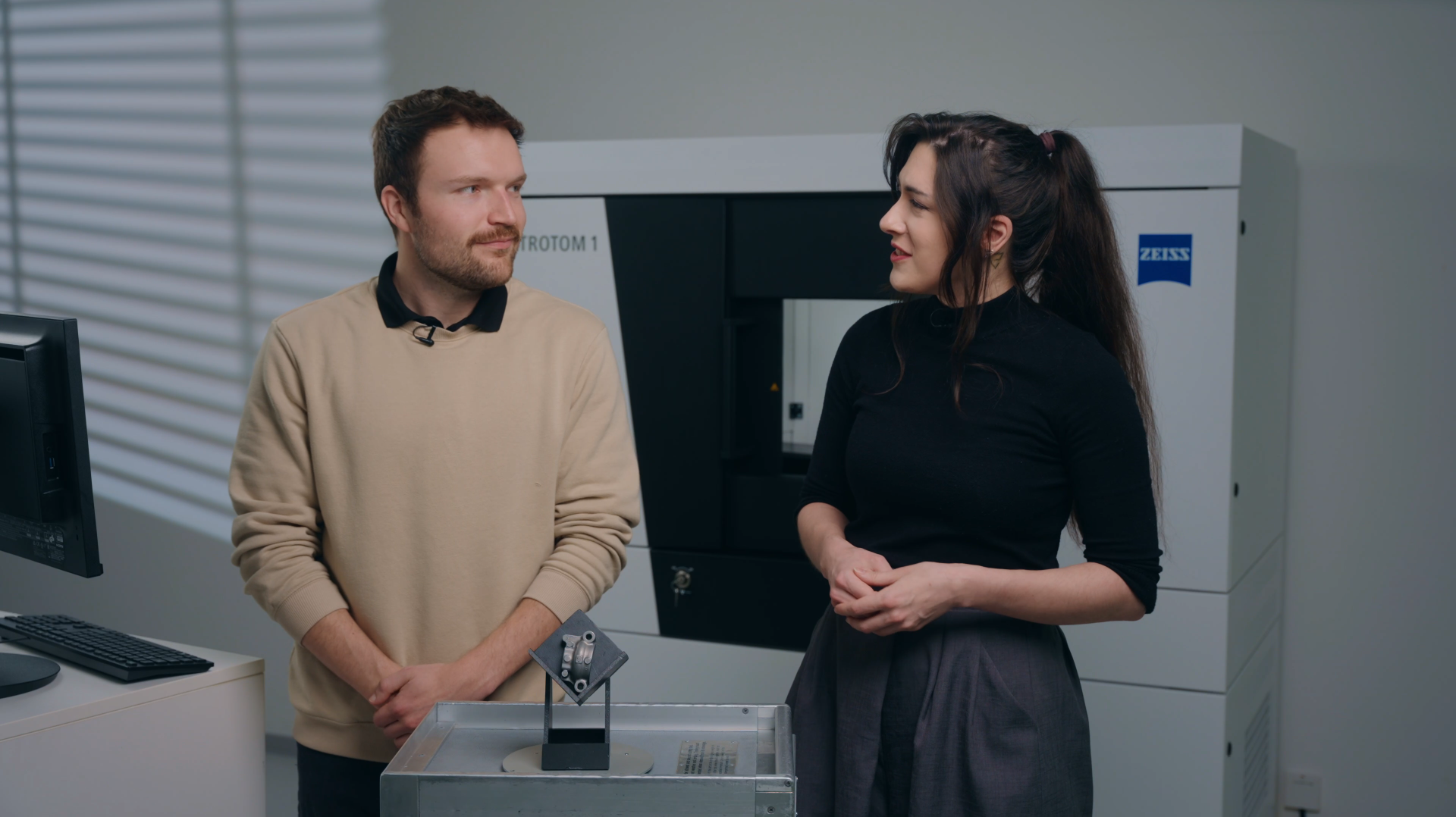A man and a woman talking about a gray part infront of a CT scanner