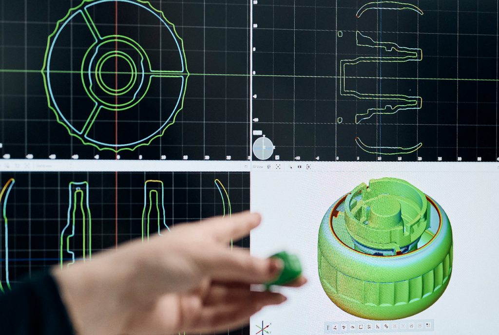 A hand with a green part in it is pointing at a software inspection of the part on a screen
